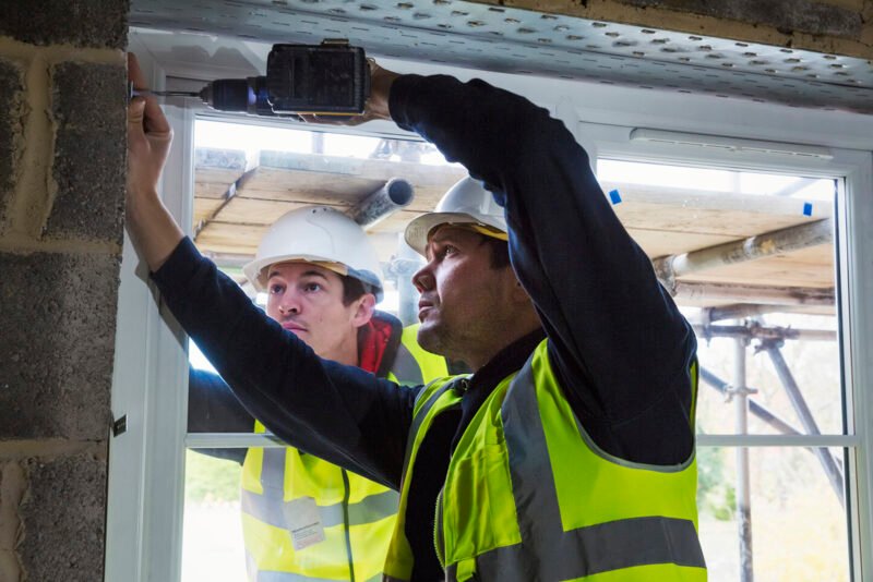 Two workmen on a construction site, builder in hard hat using an electric drill on a window frame. ,Construction Site,England